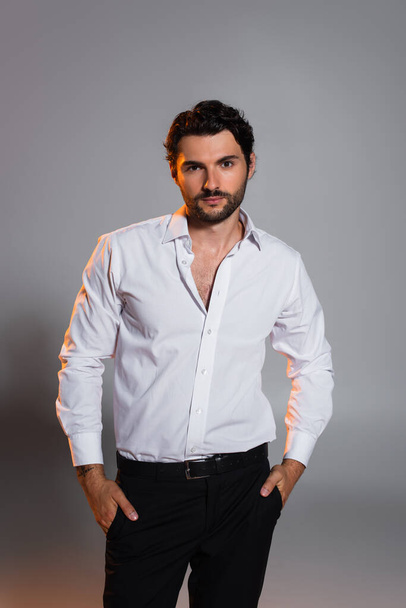stylish man in white shirt standing with hands in pockets of black trousers on grey - Photo, Image