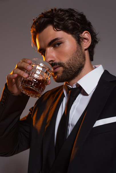 stylish man in black suit drinking whiskey while looking at camera isolated on grey - Fotografie, Obrázek