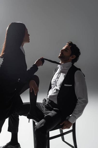 hot asian woman holding tie of elegant man sitting on chair on grey background - Fotoğraf, Görsel