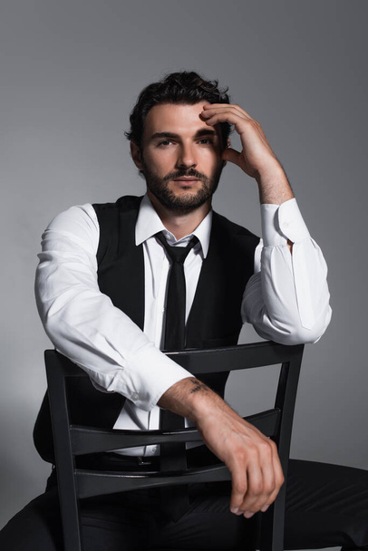elegante uomo in gilet nero e cravatta seduta sulla sedia e guardando la fotocamera isolata sul grigio - Foto, immagini