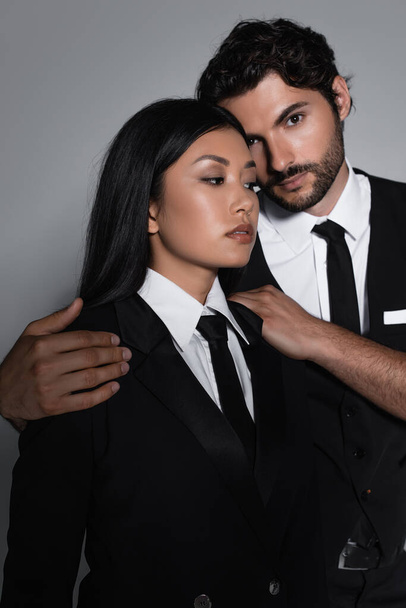 elegant brunette man looking at camera while hugging sensual asian woman isolated on grey - 写真・画像
