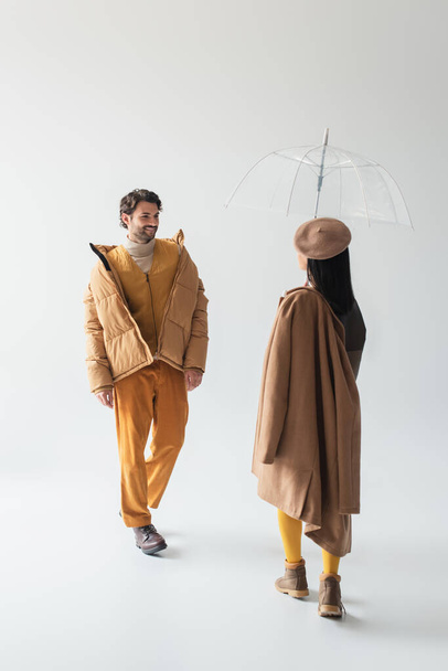 kobieta w przezroczystym parasolem i uśmiechnięty mężczyzna w beżowej kurtce buforowej podchodzący do siebie na szaro - Zdjęcie, obraz