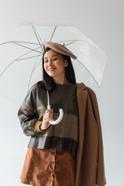 trendy asian woman standing under transparent umbrella and smiling with closed eyes isolated on grey - Photo, Image