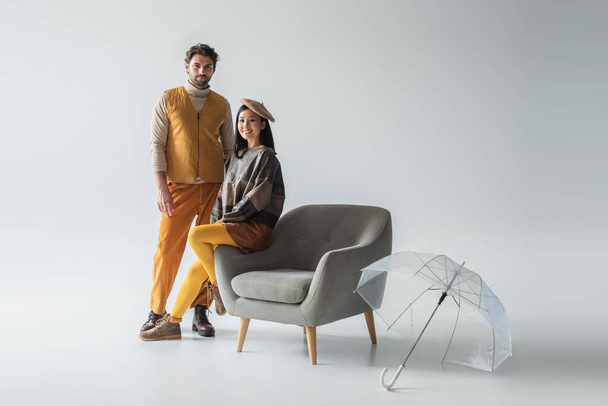 cheerful asian woman sitting on armchair near trendy man and transparent umbrella on grey - Photo, Image