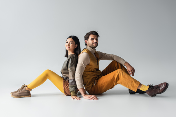 stylish interracial couple smiling at camera while sitting back to back on grey - 写真・画像