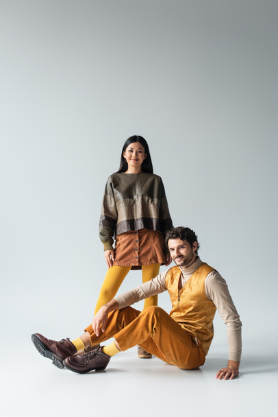 smiling man in yellow trousers sitting near asian woman in leather jacket on grey - Fotoğraf, Görsel