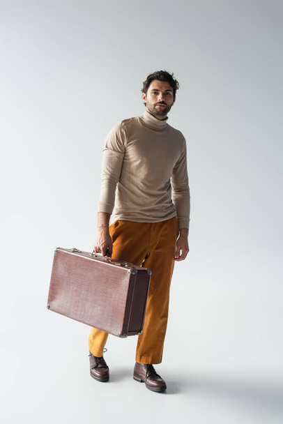 full length view of man in beige turtleneck and yellow trousers walking with vintage suitcase on grey - Φωτογραφία, εικόνα