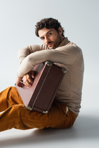 trendy man in yellow trousers and beige turtleneck sitting with vintage suitcase on grey - Photo, Image