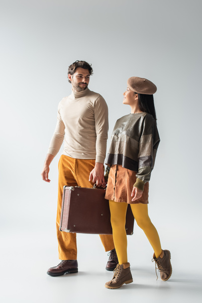 trendy interracial couple carrying vintage suitcase and smiling at each other on grey - Φωτογραφία, εικόνα