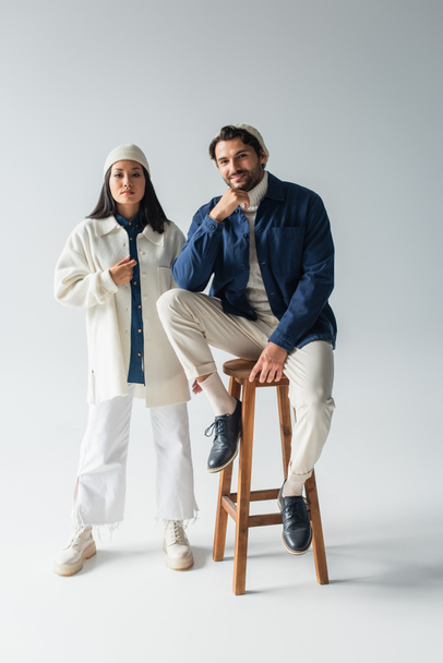 full length view of trendy asian woman near smiling man sitting on high stool on grey - Foto, afbeelding