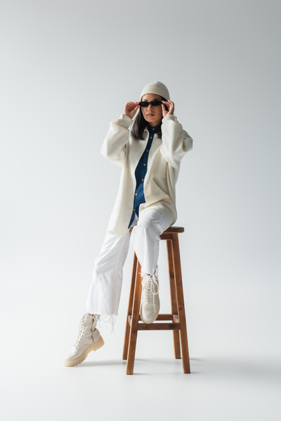 full length view of asian woman in white clothes and black sunglasses posing on high stool on grey - Foto, immagini