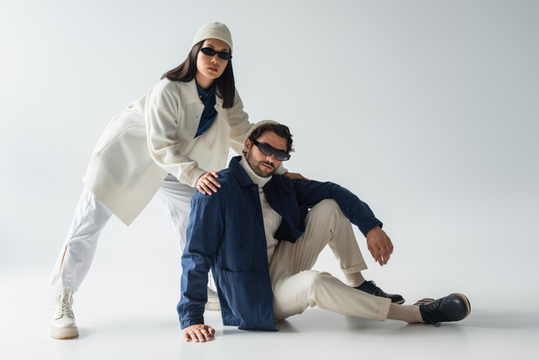 fashionable interracial couple in dark sunglasses looking at camera while posing on grey - Фото, изображение