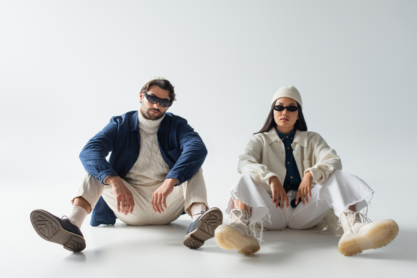 full length view of multiethnic couple in trendy clothes and dark sunglasses sitting on grey - Foto, Imagem