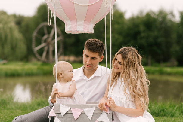 A beautiful little girl with mom and dad in a basket with a balloon and a cake celebrates her first birthday - Фото, зображення