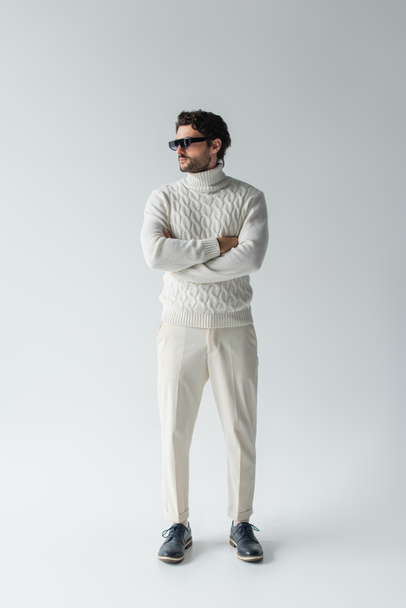 morena hombre en pantalones blancos, suéter de punto y gafas de sol oscuras de pie con los brazos cruzados en gris - Foto, Imagen