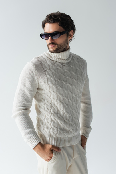 stylish man in white knitted sweater and dark sunglasses standing with hands in pockets isolated on grey - Foto, afbeelding