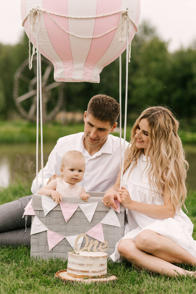 A beautiful little girl with mom and dad in a basket with a balloon and a cake celebrates her first birthday - Zdjęcie, obraz