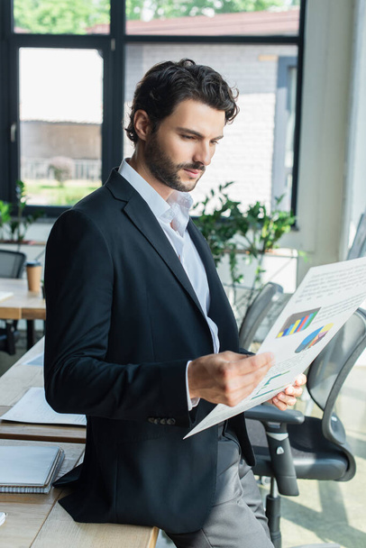bearded businessman in black blazer looking at charts while standing in office - Foto, Bild