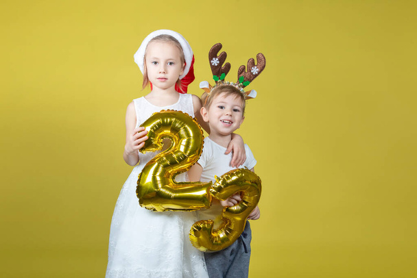 Adorables niños caucásicos en el sombrero de Santa Navidad y traje de renos sosteniendo globos con números y haciendo caras graciosas sobre un fondo amarillo. El concepto de Año Nuevo y Navidad. - Foto, imagen