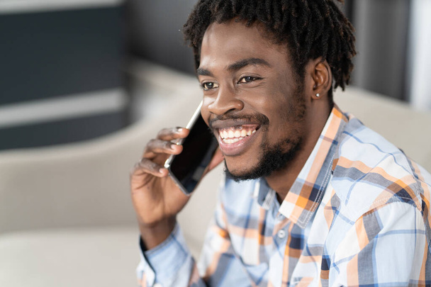 Young African American man talking on the phone holding smartphone in hand looking away. Portrait of happy man having present conversation sitting on the sofa. Social media concept - Foto, immagini