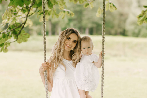Beautiful young mom with a one-year-old daughter in nature, mom and daughter riding on a swing - Foto, Imagem