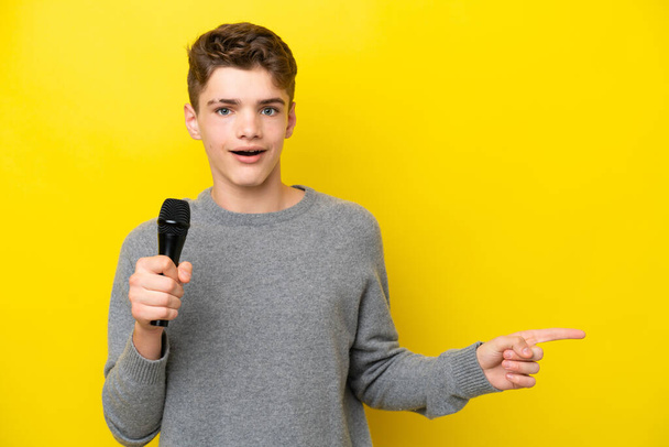 Laulaja Teini mies poimien mikrofoni eristetty keltaisella taustalla yllättynyt ja osoittaa sormella sivulle - Valokuva, kuva