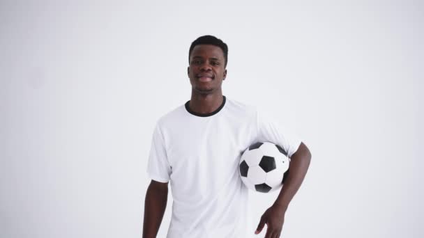 Portret van een gelukkige voetballer van een Afrikaanse etnische groep in een wit uniform op een witte achtergrond met een bal in zijn handen Lachende Afrikaanse voetballer - Video