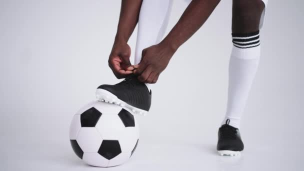 Közelkép fekete afrikai férfi focistáról, aki cipőfűzőt kötött a stúdióra, lassított felvételen. Focista bekötötte a cipőjét. Alacsony rész. összekötő labdarúgó csizma előkészítése fehér háttér - Felvétel, videó