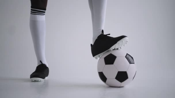 Close-up do pé de um jogador profissional de futebol preto em pé sobre a bola em câmera lenta no fundo branco. Brasileiro jogador de futebol pé na bola para posar - Filmagem, Vídeo