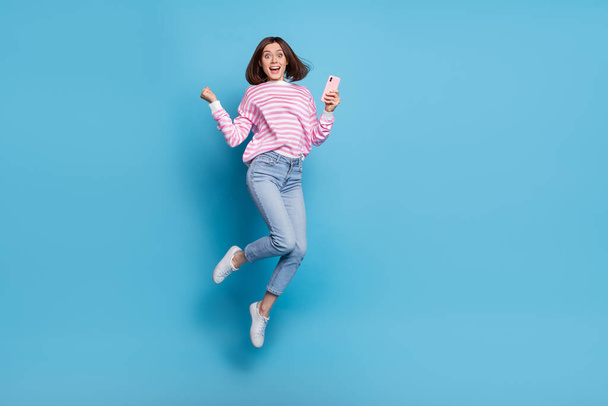 Full body photo of impresssed millennial lady jump with telephone yell wear shirt jeans sneakers isolated on blue background. - Zdjęcie, obraz