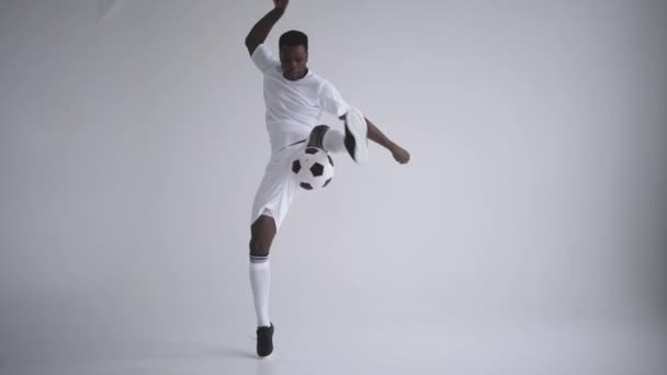 Een professionele zwarte voetballer in een wit uniform op een witte achtergrond jongleert in slow motion met een bal. Afro-Amerikaanse etnische groep voetballer met een voetbal - Video