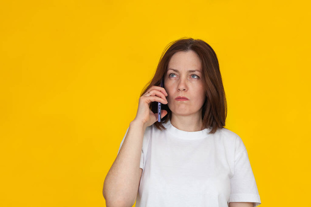 Portrait of a smiling european brunette woman wearing white T-shirt on yellow background talking on mobile phone. Business, confidence and communication concept. - Foto, Imagen