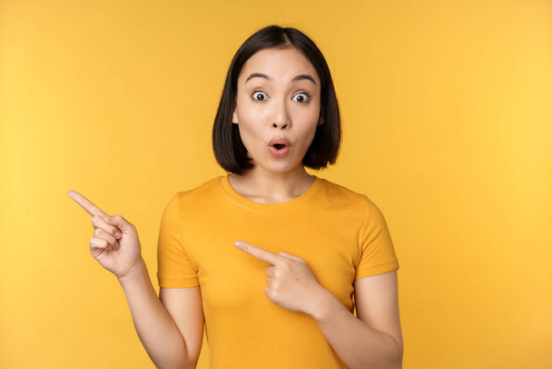 Smiling asian woman pointing fingers left, showing advertisement on empty copy space, standing over yellow background - Foto, afbeelding