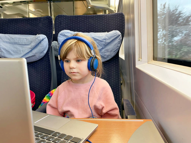 かわいい就学前の女の子は電車に座って、移動中にラップトップでテレビを見ています。鉄道で家族旅行に行く幸せな子供。ノートとヘッドフォンを持つ女の子 - 写真・画像