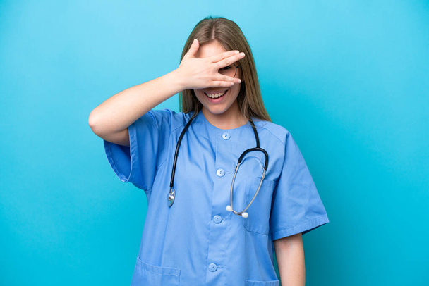 Νεαρή χειρουργός γιατρός γυναίκα απομονωμένη σε μπλε φόντο που καλύπτει τα μάτια με τα χέρια και χαμογελά - Φωτογραφία, εικόνα
