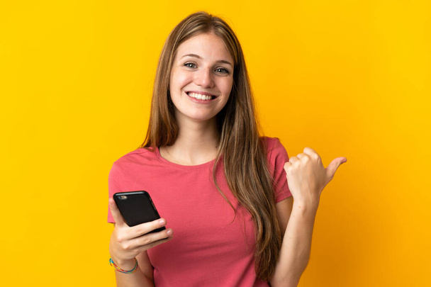 Jonge vrouw met behulp van mobiele telefoon geïsoleerd op gele achtergrond wijzend naar de zijkant om een product te presenteren - Foto, afbeelding