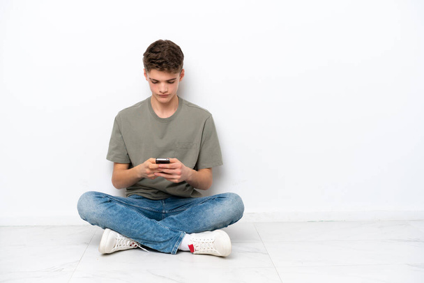 Genç Rus adam, beyaz arka planda izole bir şekilde yerde oturuyor ve cep telefonuyla bir mesaj gönderiyor. - Fotoğraf, Görsel