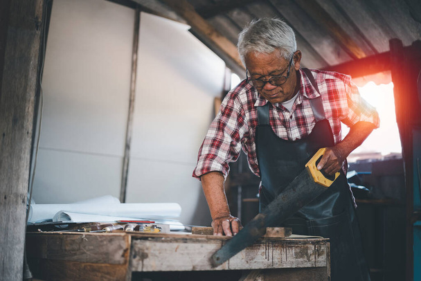 Senior old carpenter using saw working on wood craft at workshop to produce construction material or wooden furniture. - Photo, Image