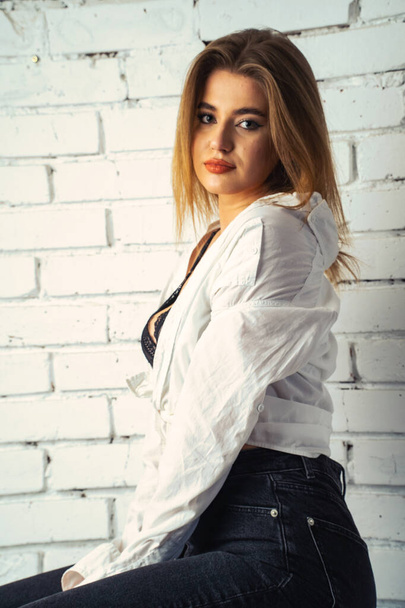 Фото молодой красивой чувственной и сексуальной женщины с длинными волосами в белой рубашке, позирующей против белой кирпичной стены в студии.   - Фото, изображение