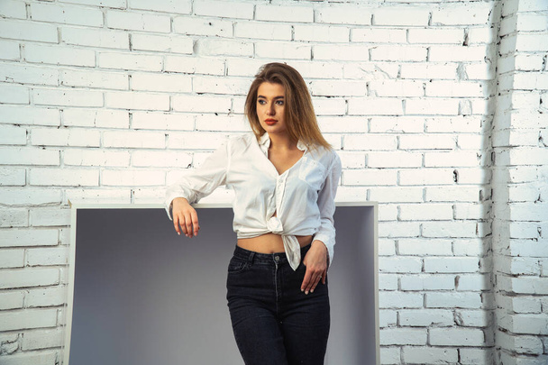 Photo of young beautiful sensual and sexy woman with long hair wearing in white shirt posing against a white brick wall in the studio.   - Zdjęcie, obraz