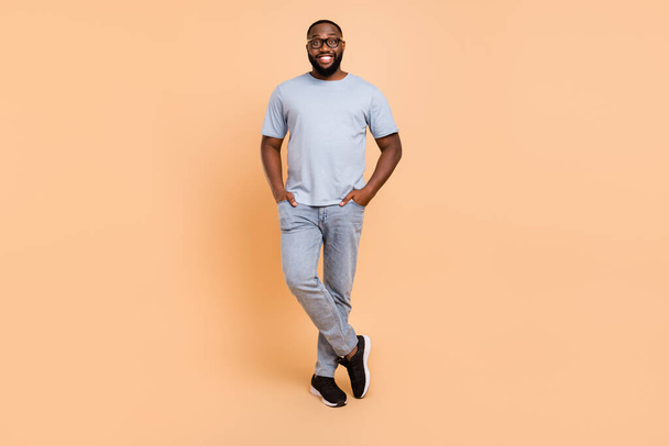 Full size photo of nice young beard guy stand wear eyewear blue t-shirt jeans sneakers isolated on beige color background - Fotoğraf, Görsel