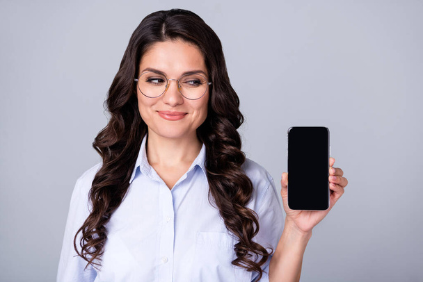 Fénykép az érdeklődő hölgy tartsa telefon nézd oldalán üres tér képernyő kopás szemüveg kék ing elszigetelt szürke szín háttér - Fotó, kép