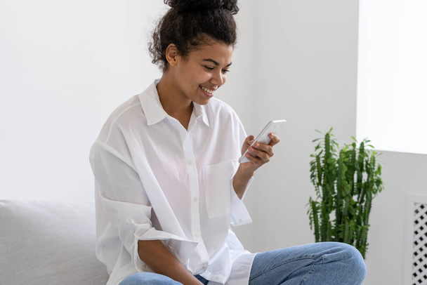 Retrato de una linda chica afro americana sonriente charlando con amigos usando un teléfono inteligente. Comunicación en línea. Concepto de adicción a los aparatos. Estilo de vida de los adolescentes - Foto, Imagen