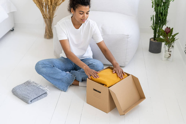 High above view of afro american teenager folds clothes into box. Minimalism lifestyle. Process of moving to another apartment. Things charity concept - 写真・画像