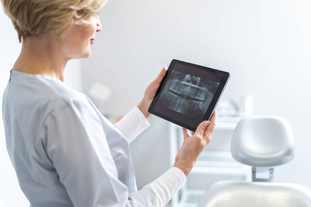 Ciddi profesyonel dişçi hastasının diş röntgenleriyle dijital tableti tutuyor, radyografi fotoğraflarına bakıyor, stomatoloji kliniğinde duruyor. Tedavi, protez, diş çıkarma konsepti - Fotoğraf, Görsel