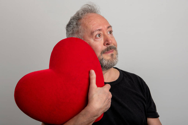 Un bel homme barbu et aux cheveux gris d'âge moyen tenant un cœur rouge, un fond gris isolé. Concept de Saint-Valentin - Photo, image