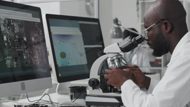 Ingeniero masculino afroamericano en bata de laboratorio examinando microprocesador chip bajo microscopio y mirando pantallas de computadora mientras trabaja con electrónica - Metraje, vídeo