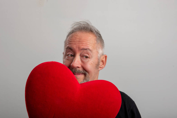 Hombre guapo de mediana edad de pelo gris y barbudo que sostiene un corazón rojo, fondo gris aislado. Concepto de San Valentín - Foto, Imagen