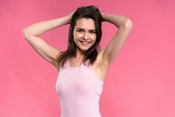 Young brunette woman in sleeveless t-shirt tighten hair back head with hands up isolated on pink background - Foto, Imagen