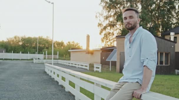 Mittellanger blauäugiger kaukasischer Mann in lässiger Kleidung, der sich im Sommer abends in der Nachbarschaft an einen weißen Zaun lehnt, lächelt und in die Kamera blickt - Filmmaterial, Video